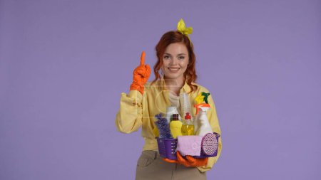 Photo for Everyday cleaning and housekeeping concept. Woman in casual clothing and rubber gloves holding basket with cleaners and rugs, holding hand up pointing. Isolated on purple background. - Royalty Free Image