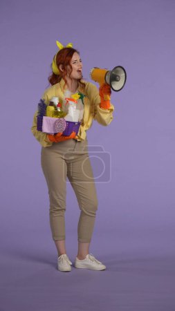 Photo for Everyday housekeeping creative concept. Vertical photo. Woman in casual clothing and rubber gloves holding basket with cleansers and shouting in megaphone. - Royalty Free Image