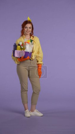 Photo for Everyday housekeeping creative concept. Vertical photo. Woman in casual clothing and rubber gloves smiling holding basket with cleansers, posing at the camera. Space for advertisement. - Royalty Free Image