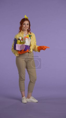 Photo for Everyday housekeeping creative concept. Vertical photo. Woman in casual clothing and rubber gloves smiling holding basket with cleansers showing empty area. Space for advertisement. - Royalty Free Image