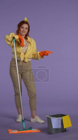 Photo for Everyday housekeeping creative concept. Vertical photo. Woman in casual clothing and rubber gloves holding mop, pointing at the empty area. Space for advertisement. - Royalty Free Image
