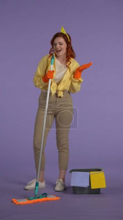 Photo for Everyday housekeeping creative concept. Vertical photo. Woman in casual clothing and rubber gloves mopping the floor with a mop, singing song, happy face expression. - Royalty Free Image