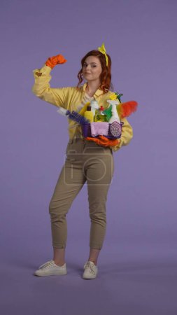 Photo for Everyday housekeeping creative concept. Vertical photo. Woman in casual clothing and rubber gloves with smiling face holding basket with cleansers, showing strong hand gesture. - Royalty Free Image