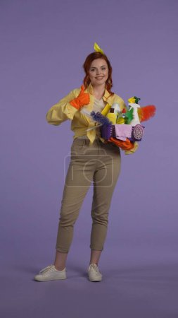 Photo for Everyday housekeeping creative concept. Vertical photo. Woman in casual clothing and rubber gloves with smiling face holding basket with cleansers, showing thumbs up. - Royalty Free Image