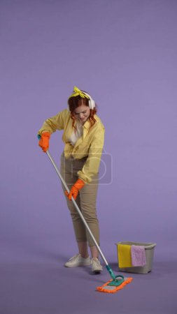 Photo for Everyday housekeeping creative concept. Vertical photo. Woman in casual clothing and rubber gloves wearing headphones listening music and mopping the floor. - Royalty Free Image