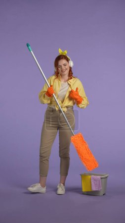 Photo for Everyday housekeeping creative concept. Vertical photo. Woman in casual clothing and rubber gloves in headphones listening music, holding mop, shows thumbs up. - Royalty Free Image