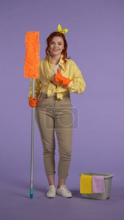 Photo for Everyday housekeeping creative concept. Vertical photo. Woman in casual clothing and rubber gloves standing with mop, showing thumbs up smiling looking at the camera. - Royalty Free Image