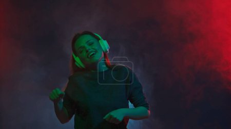 Foto de Una joven pelirroja con auriculares inalámbricos blancos escucha música y bailes de DJ. Una mujer baila en un estudio en nubes de humo con luces de neón multicolores - Imagen libre de derechos