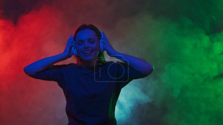 Foto de Una joven pelirroja con auriculares inalámbricos blancos escucha música y bailes de DJ. Una mujer baila en un estudio en nubes de humo con luces de neón multicolores - Imagen libre de derechos