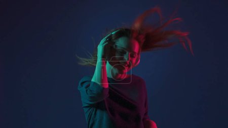 Photo for A young redhaired woman wearing white wireless headphones listens to DJ music and dances. Woman in studio on blue background in pink and green neon light - Royalty Free Image