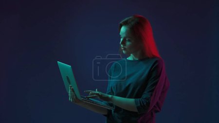 Photo for A woman works on a laptop, types a text, message, communicates. Woman with laptop in hand in studio on blue background in pink and green neon light. Remote work, freelancer - Royalty Free Image