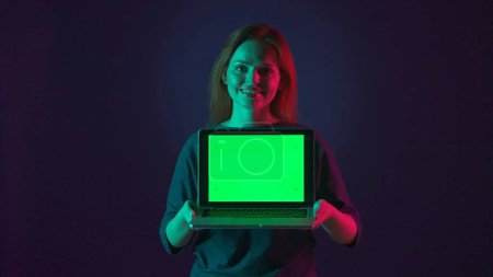 Téléchargez les photos : Femme montrant écran vert ordinateur portable de près. Portrait d'une femme avec un ordinateur portable avec un écran vert dans le studio sur un fond bleu en néon rose et vert. Espace publicitaire, lieu de travail - en image libre de droit