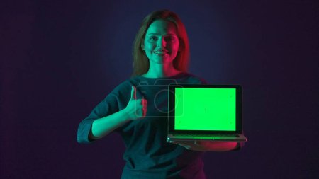 Téléchargez les photos : Une femme tient un ordinateur portable avec un écran vert et montre les pouces vers le haut. Portrait d'une femme avec un ordinateur portable avec un écran vert en studio sur fond bleu en néon rose et vert - en image libre de droit
