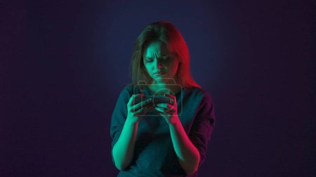 Téléchargez les photos : Portrait d'une femme déçue qui a perdu dans un jeu en ligne sur un smartphone. Femme en studio sur fond bleu en néon rose et vert - en image libre de droit