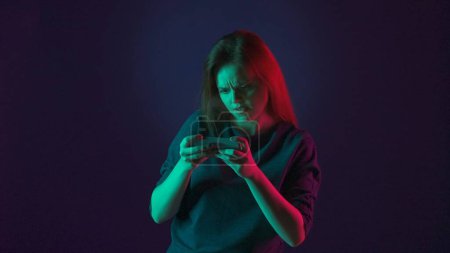 Foto de Una mujer tensa juega un juego en línea en su teléfono inteligente. Mujer en estudio sobre fondo azul en luz de neón rosa y verde - Imagen libre de derechos