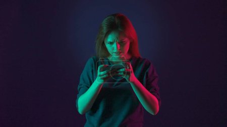 Téléchargez les photos : Une femme tendue joue à un jeu en ligne sur son smartphone. Femme en studio sur fond bleu en néon rose et vert - en image libre de droit