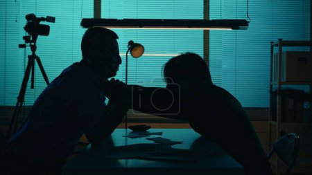 Photo for Medium silhouette shot of a perpetrator or prisoner sitting in the interrogation room with the detective. Suspect tries to strangle him. True crime, documentary, creative content. - Royalty Free Image