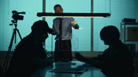 Photo for Silhouette, medium shot of a suspect, perpetrator, prisoner sitting in the interrogation room with detective and policeman. They try to get the confession. True crime, documentary, creative content. - Royalty Free Image