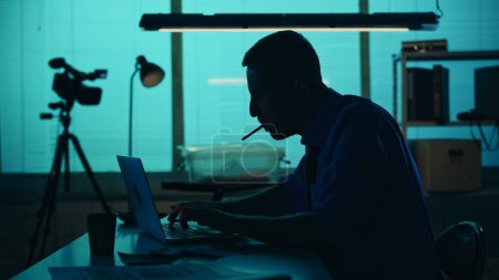 Photo for Medium silhouette shot of a detective, policeman sitting in the interrogation room, smoking, working on a laptop and studying the evidence. True crime, documentary, creative content. - Royalty Free Image