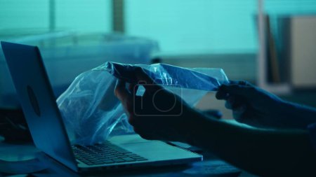 Photo for Close up detail silhouette shot of a detective, policeman sitting in the interrogation room, working on a laptop and studying a knife from the evidence. True crime, documentary, creative content. - Royalty Free Image