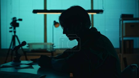 Photo for Medium silhouette shot of a suspect, offender, perpetrator or prisoner sitting in the interrogation room. He is studying the evidence on the table. True crime, documentary, creative content. - Royalty Free Image