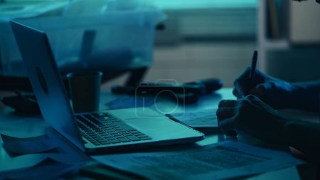 Photo for Close up detail silhouette shot of a detective, policeman sitting in the interrogation room, working on a laptop and studying the evidence. True crime, documentary, creative content. - Royalty Free Image