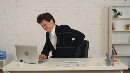 Photo for Medium shot of a young man sitting at the table, working on a laptop, massaging, warming up as if his back is hurting after a long day, shift. Overworking, health issues, medical, sedentary lifestyle. - Royalty Free Image