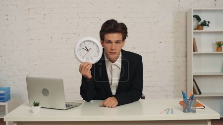 Foto de Mediana toma de un joven sentado en la mesa de la oficina, sosteniendo un reloj en la mano, mirando a la cámara, soñoliento, cansado y agotado. Negocios, exceso de trabajo, sedentarismo. - Imagen libre de derechos