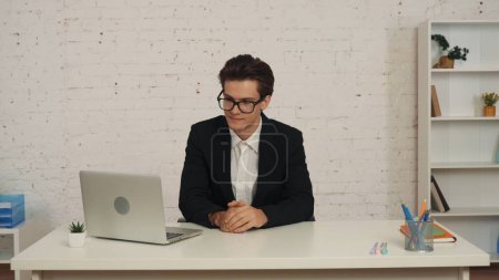 Téléchargez les photos : Plan moyen d'un jeune homme d'affaires portant des lunettes, assis à la table dans un bureau, travaillant sur son ordinateur portable, concentré, consentant et heureux. Troubles, ophtalmologie, publicité pour les services médicaux. - en image libre de droit