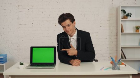 Photo for Medium shot of a young man in an office, sitting at the table with a laptop that has an advertising area, workspace mock up on it. The man points at the screen. Business, web resources, app. - Royalty Free Image
