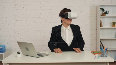Téléchargez les photos : Plan moyen d'un jeune homme dans un bureau portant la réalité virtuelle, casque vr, équipement, regardant, interagissant avec un écran invisible excité et heureux. Technologie moderne, effets visuels, opportunités. - en image libre de droit