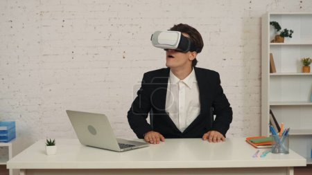 Foto de Fotografía media de un joven en una oficina vistiendo realidad virtual, casco vr, equipo, mirando, interactuando con una pantalla invisible emocionado y feliz. Tecnología moderna, efectos visuales, oportunidades. - Imagen libre de derechos