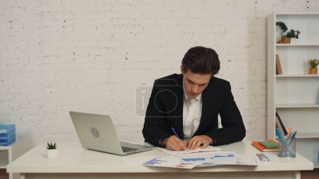 Photo for Medium shot of a young man sitting in the office, studying and cheching the graphs and charts on his table. He takes notes, writes something down. Business advertisement, official style. - Royalty Free Image