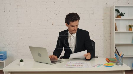 Téléchargez les photos : Plan moyen d'un jeune homme assis dans le bureau, travaillant sur un ordinateur portable et reprenant les graphiques et les graphiques sur sa table. Il a l'air concentré. Publicité commerciale, emploi, recherche. - en image libre de droit