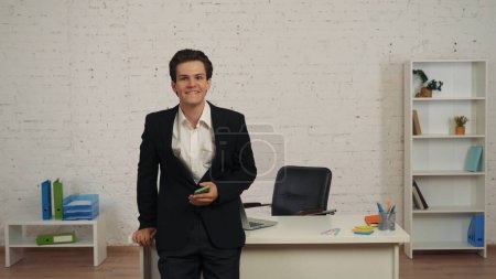 Photo for Medium shot of a young man in an office, holding a smartphone in hand, receiving a message, notification with good news, offer. He reacts with joy and excitement. Job opportunity, advertisement. - Royalty Free Image