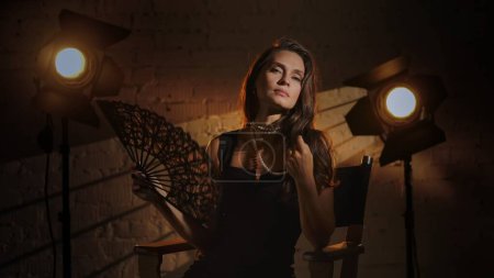 Photo for Cinematography and movie backstage advertisement creative concept. Brunette woman in black dress and pearl necklace sitting on a directors chair holding opened lace fan, looking at the camera. - Royalty Free Image