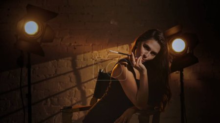 Photo for Cinematography and movie backstage advertisement creative concept. Woman in black dress and pearl necklace sitting on a directors chair, holding cigarette holder and smoking, posing at the cam. - Royalty Free Image