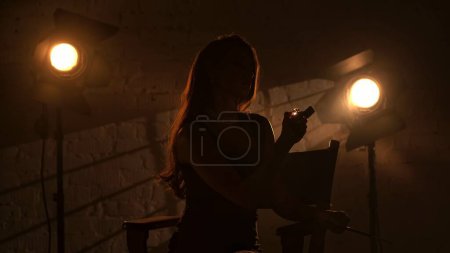 Foto de Cinematografía y cine detrás del escenario publicidad concepto creativo. Mujer en vestido negro y collar de perlas sentado en una silla de directores, sosteniendo el sostenedor del cigarrillo y trata de iluminar el encendedor. - Imagen libre de derechos