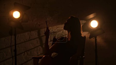Foto de Cinematografía y cine detrás del escenario publicidad concepto creativo. Hermosa silueta de mujer en vestido negro sentado en una silla de directores, sosteniendo elegantemente el sostenedor del cigarrillo y fumando. - Imagen libre de derechos