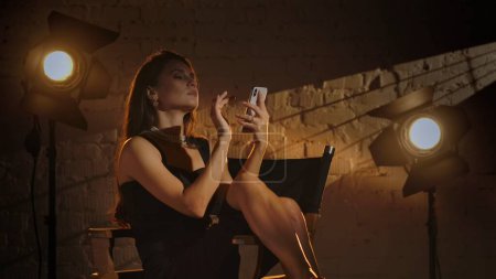 Foto de Cinematografía y cine detrás del escenario publicidad concepto creativo. Mujer atractiva en vestido negro y collar de perlas sentado en una silla de directores, sosteniendo y escribiendo mensaje en el teléfono inteligente. - Imagen libre de derechos