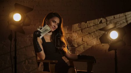 Photo for Cinematography and movie backstage advertisement creative concept. Woman in black dress and pearls in silk gloves sitting on a directors chair, holding wad of money bills and smiling at the camera. - Royalty Free Image