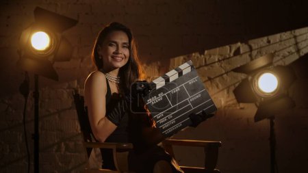 Photo for Cinematography and movie backstage advertisement creative concept. Beautiful woman in black dress, pearls and silk gloves sitting on a directors chair, holding clapperboard and smiling at the camera. - Royalty Free Image