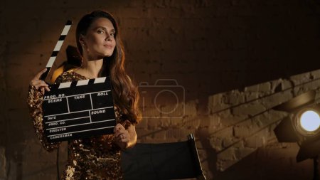 Photo for Cinematography and movie backstage advertisement creative concept. Closeup shot of beautiful woman in golden dress, standing next to directors chair, holding clapperboard and looking at the camera. - Royalty Free Image