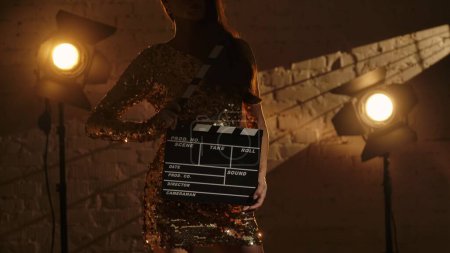 Téléchargez les photos : Cinématographie et film publicité dans les coulisses concept créatif. Gros plan de la silhouette de la femme brune en robe dorée, debout dans le studio contre le mur de briques et tenant clapperboard. - en image libre de droit