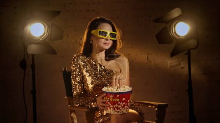 Photo for Cinematography and movie backstage advertisement creative concept. Closeup shot of brunette woman in golden dress, sitting on directors chair, in paper glasses and eating popcorn from a bucket. - Royalty Free Image