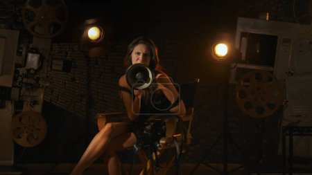 Photo for Cinematography and movie backstage advertisement creative concept. Brunette in black dress sitting on directors chair behind the scene, holding megaphone and shouting something looking at the camera. - Royalty Free Image