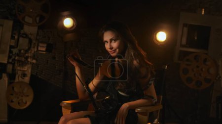 Téléchargez les photos : Cinématographie et film publicité dans les coulisses concept créatif. Femme attrayante en robe noire assise sur la chaise des réalisateurs dans les coulisses, souriant tenant des bandes de film, regardant la caméra. - en image libre de droit