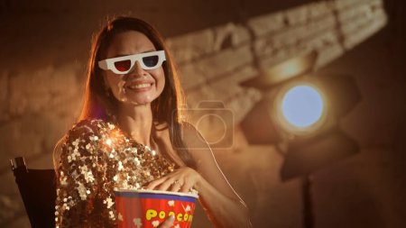 Photo for Cinematography and movie backstage. Closeup shot of beautiful brunette woman actress in golden dress sitting on directors chair and watching comedy, wearing 3d glasses eating popcorn. - Royalty Free Image