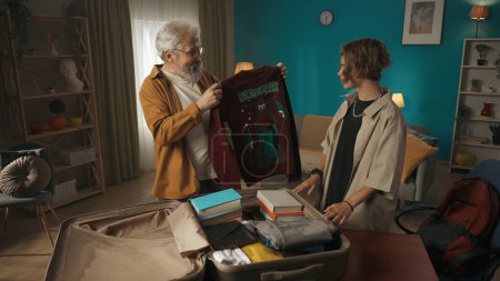 Photo for Shot of a teenager, young man packing his belongings in a suitcase. His grandfather is helping him, offering him to take a sweater. College, university, moving to a new place. - Royalty Free Image