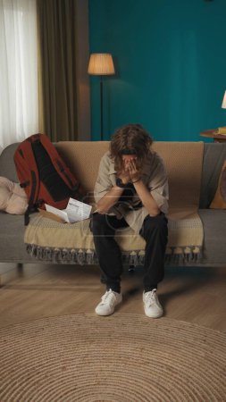Photo for Vertical shot of a teenager, young man sitting on a sofa, couch, devastated and frustrated about his fail. There is a document, denied admission paper next to him, College, university, enrollment. - Royalty Free Image